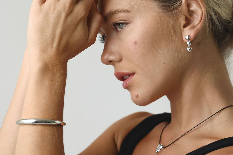 A blonde, blue eyed woman, with her profile to camera, and hands up near forehead, wears silver heart shaped earrings, a silver bangle cuff and a silver heart rope necklace