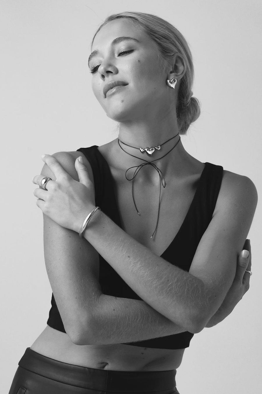 Black and white image of woman with blonde low bun, wearing a black crop top and black pants, wrapping her arms around herself, showcasing her silver jewellery. A cuff, a ring, a rope necklace and heart earrings