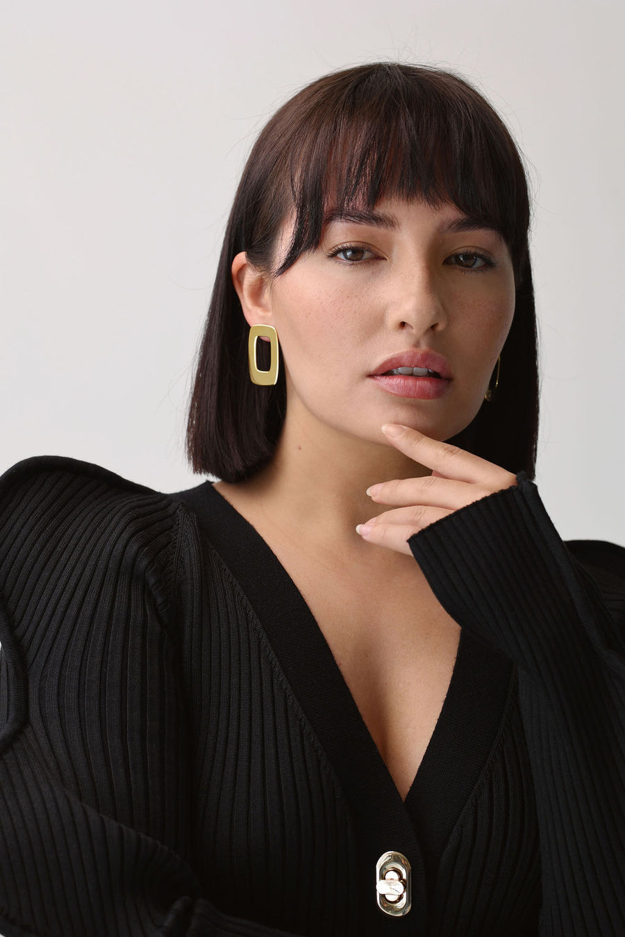 Woman with bob length brown hair and a fringe, wearing bold gold earrings, a black knit top, looking directly into the camera, touching her left finger to her chin
