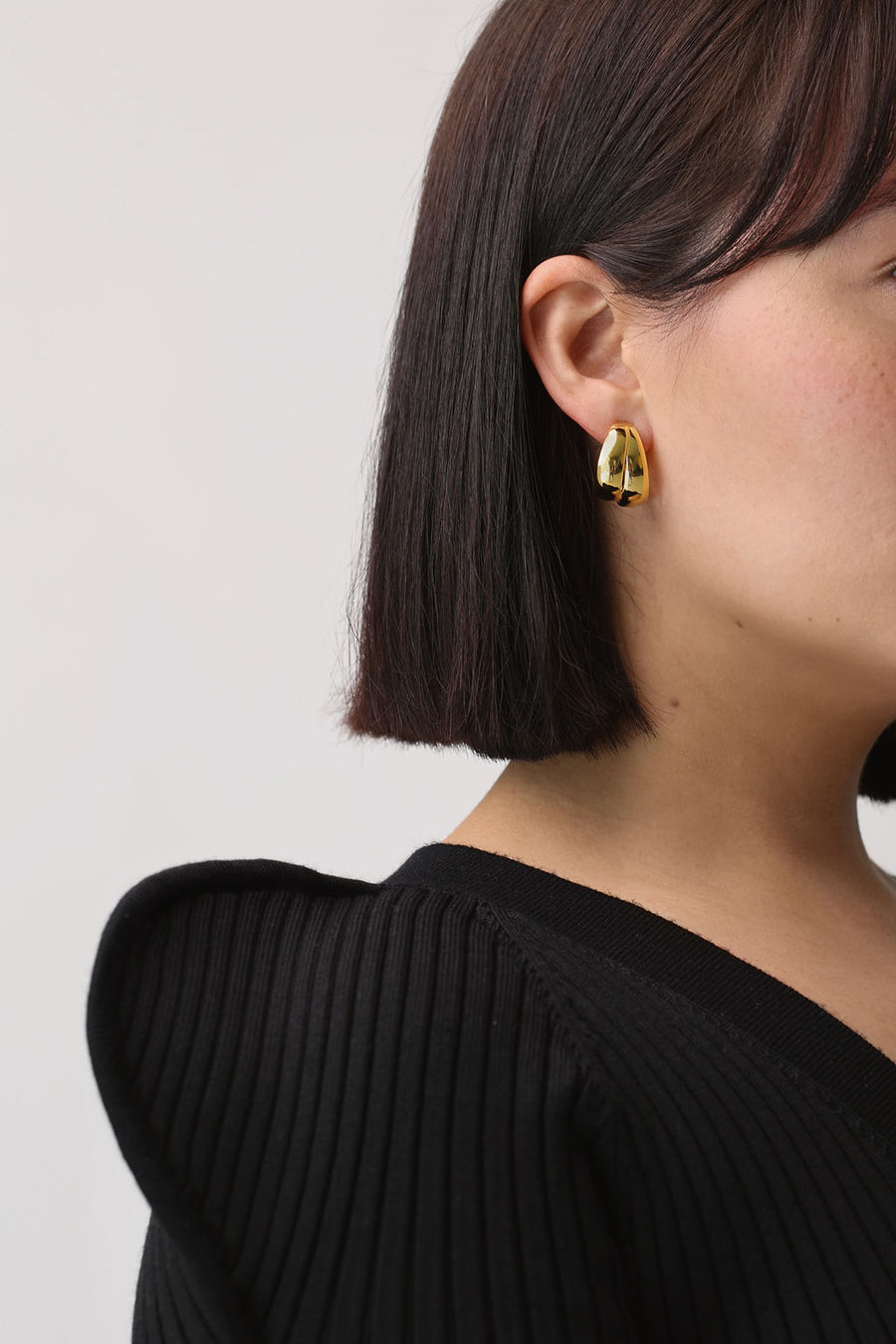 Close up of gold earring on woman's ear. She has bob length brown hair and is wearing a black knit 