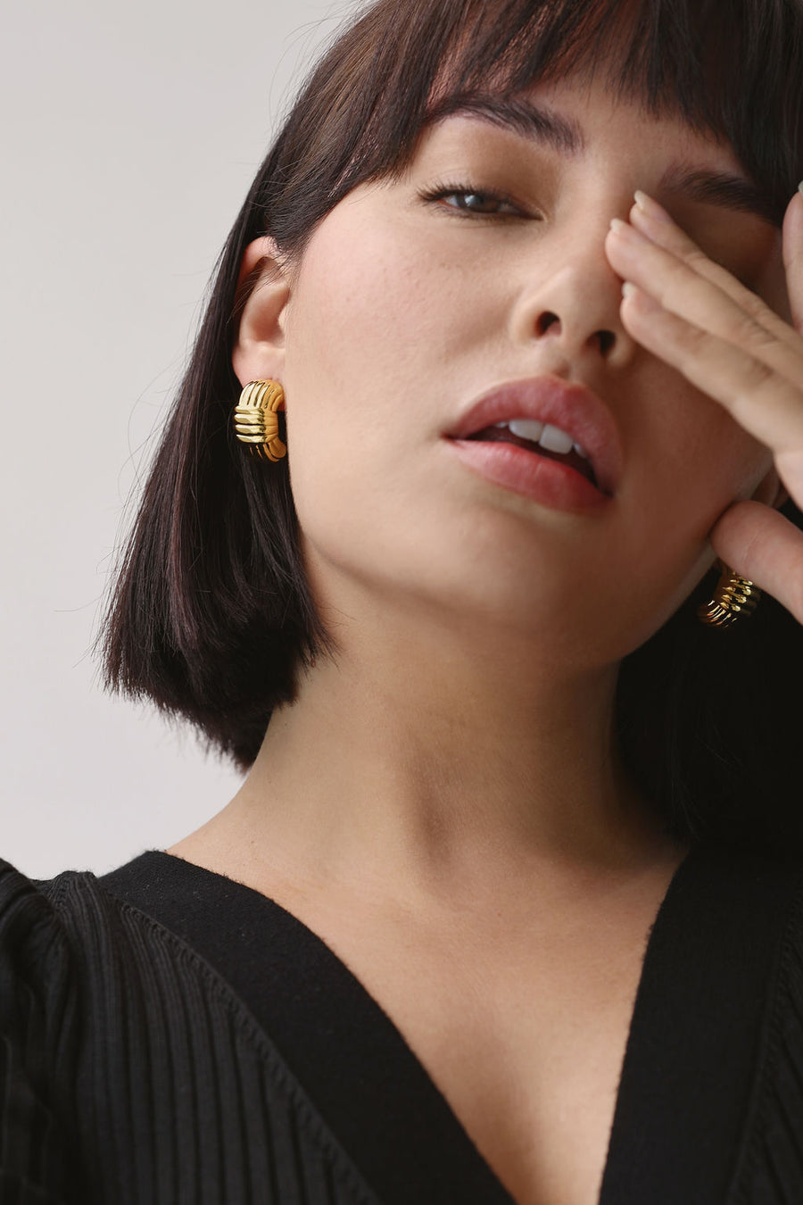 woman with bob length brown hair, weaing chunky gold earrings, staring down the camera, covering her left eye with her fingers