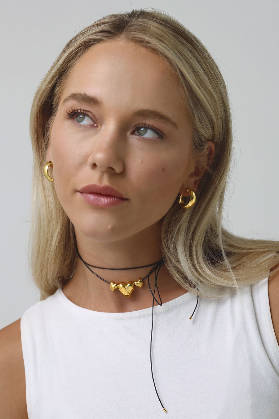 A blonde woman, looking off to her right, wearing a white top, gold hoop earrings and a wrap necklace with gold heart pendants